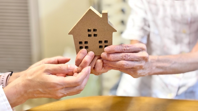 高齢者から若い世代へ木製の家の模型を手渡す不動産相続のイメージ