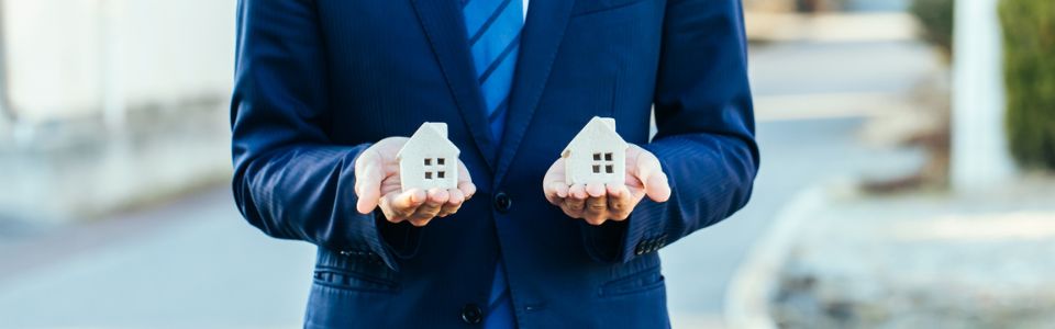 両手に大小の家の模型を持つスーツ姿の不動産営業担当者