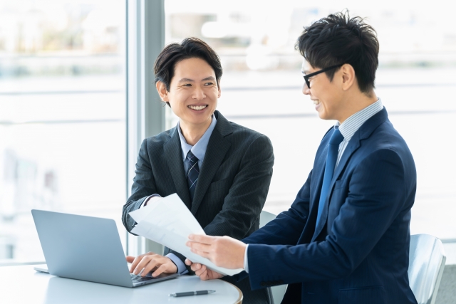 ノートパソコンと書類を前に笑顔で打ち合わせをする2人のビジネスマン